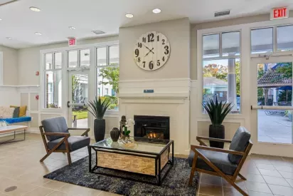 A welcoming seating area with a fireplace, large clock, and natural light from floor-to-ceiling windows.