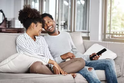 A cheerful couple sitting on a cozy beige sofa, sharing a laugh and enjoying a moment together in a bright and airy living room with large windows that let in natural light.