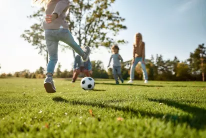 Children and adults playing soccer on a grassy field under a sunny blue sky.