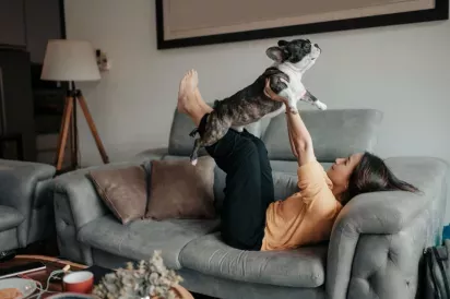 A woman lays on the couch while holding her dog in the air like Superman