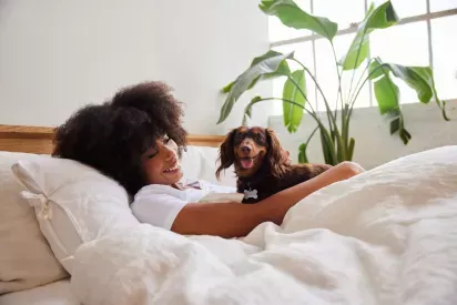 A woman lays in bed while smiling at her dog who is smiling at the camera