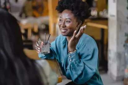 A woman in a teal blouse sits at a table in a casual setting, holding a cocktail with a garnish while smiling and gesturing as she engages in conversation with someone across from her.