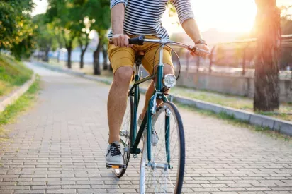 A cyclist wearing a striped shirt and yellow shorts rides a vintage-style bicycle on a tree-lined brick path, illuminated by the warm glow of the setting sun.