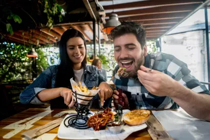 A smiling couple enjoys a meal at an outdoor restaurant, with the man eagerly taking a bite of barbecue and the woman reaching for fries from a stylish spiral holder.