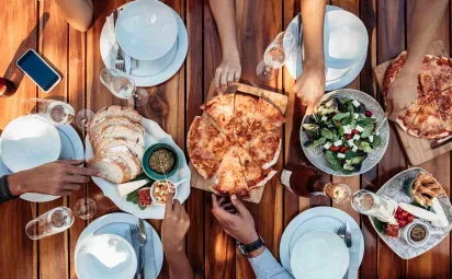 A top-down view of a wooden table set with pizza, salad, bread, and appetizers, surrounded by people sharing the meal.