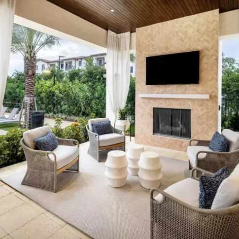 A luxurious covered seating area featuring a built-in fireplace, a mounted TV, and modern wicker chairs with blue cushions, surrounded by lush landscaping.