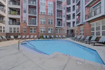 An outdoor swimming pool surrounded by lounge chairs, framed by a multi-story apartment building with balconies.