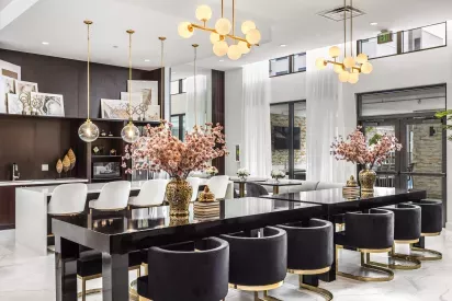 A luxurious communal space featuring a black dining table with velvet chairs, elegant gold chandeliers, and large windows with natural light.