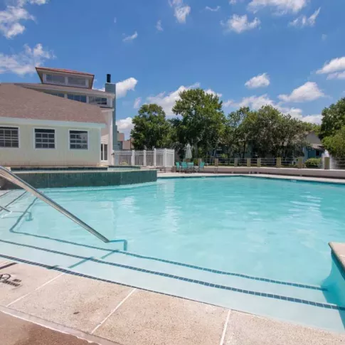 A sparkling outdoor pool with a stair entry, surrounded by a sunny deck, a poolside building, and lush trees under a bright blue sky.