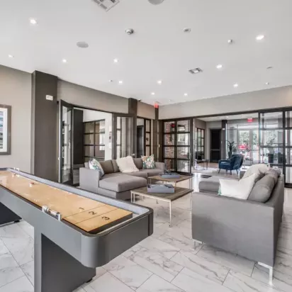 A lounge space with comfortable seating, a shuffleboard table, and glass-paneled doors.