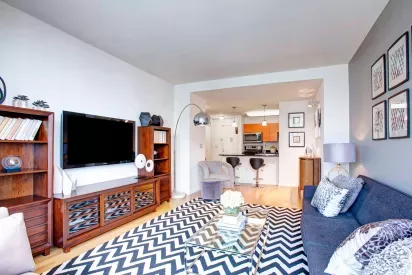 A modern living room with large windows offering city views, stylish furniture, and a bold zigzag patterned rug, creating a vibrant and cozy ambiance.