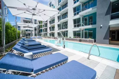 "Resort-style pool area at The Pacific Luxury Apartments featuring modern blue lounge chairs, large white umbrellas, and sleek apartment balconies with glass railings."