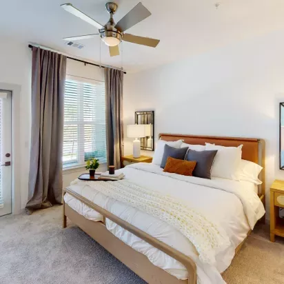 Spacious bedroom with a modern design, featuring a plush bed with layered pillows, bedside tables, and soft natural light through large windows.