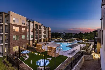 An aerial view of The Warner Apartments' pool and courtyard at dusk, featuring glowing string lights, shaded lounge areas, and a well-maintained green space.