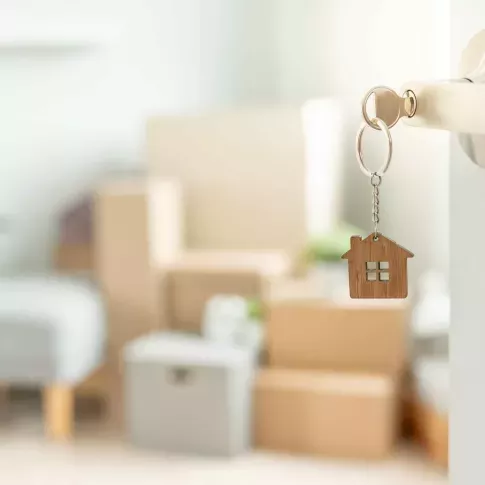 Close-up of a silver door handle with a key inserted, featuring a wooden house-shaped keychain. In the blurred background, a bright and modern living space is filled with moving boxes, a gray ottoman, and green plants, symbolizing a new home or apartment move-in.