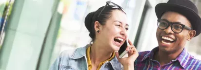 A cheerful young woman laughs while talking on the phone, accompanied by a smiling man in stylish glasses and a hat.