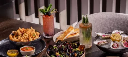 A table spread featuring gourmet dishes, including crispy calamari with dipping sauce, herb-garnished mussels, oysters on ice with vibrant garnishes and lemon, toasted bread slices, and two cocktails with decorative garnishes.