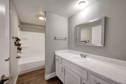 Modern bathroom featuring a long speckled white vanity, framed mirror, and a combination bathtub and shower with neutral-toned walls and wood-style flooring.