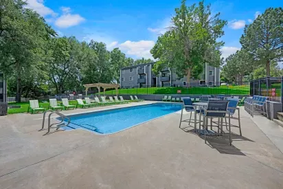 Outdoor swimming pool area featuring green-striped lounge chairs, shaded pergola seating, and modern blue patio furniture, surrounded by mature trees and grey apartment buildings.