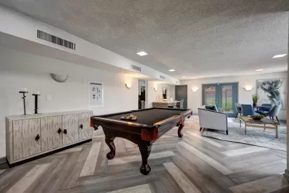 A modern community game and lounge room featuring a dark wood pool table with a black felt surface and orange tassels. The room has contemporary decor, including a textured sideboard with geometric patterns, a seating area with a gray couch and blue chairs, and a round dining table. The flooring is designed in a chevron pattern with shades of gray and beige. Large glass doors in the background lead to an outdoor area. ​​