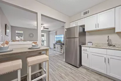 Bright, modern kitchen with white cabinetry, granite countertops, and stainless steel appliances, opening to a cozy dining nook with a wooden table and upholstered stools, and a living room with a ceiling fan and wall-mounted TV in view.