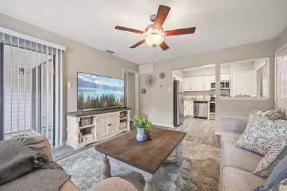Cozy living room with a ceiling fan, neutral-toned decor, flat-screen TV, and view into a modern kitchen featuring stainless steel appliances and white cabinetry.