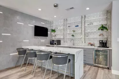 Contemporary resident lounge with a sleek white waterfall island, modern barstools, geometric accent wall, open shelving with decor, and built-in appliances including a beverage fridge and coffee station.