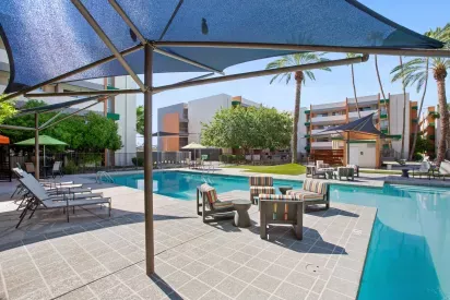 Resort-style pool area featuring modern lounge chairs, shaded seating with striped cushions, and large triangular canopies providing sun protection, surrounded by palm trees and contemporary apartment buildings.