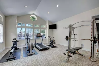 Modern fitness center with cardio and strength training equipment including a rowing machine, elliptical, treadmill, stationary bike, and a Hoist Smith machine. The room features tall ceilings, large windows allowing natural light, and carpeted flooring for comfort. "HarborFit" branding is visible on the wall.