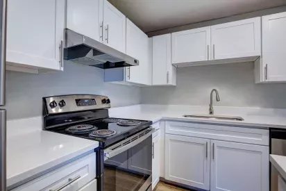 Contemporary kitchen with sleek white cabinetry, stainless steel appliances, and an electric stovetop. The space features modern brushed nickel hardware, quartz-style countertops, and a gooseneck faucet above a deep single-basin sink. The neutral gray backsplash and clean design offer a bright and minimalist aesthetic.