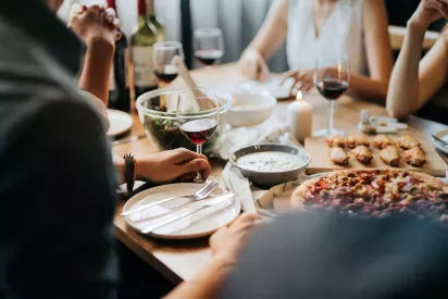 A group of friends gathered around a wooden dining table, enjoying a meal with pizza, salad, and glasses of red wine.