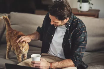 A man sitting on a couch with a cup of coffee, gently petting an orange tabby cat that is standing beside him.