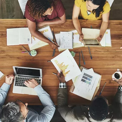 Top-down view of a diverse group of professionals collaborating around a wooden table in a modern office setting. They are analyzing data, discussing reports with charts and graphs, using a laptop and a tablet, and taking notes. The workspace is filled with pens, notebooks, and small potted plants, reflecting a dynamic and productive teamwork environment.