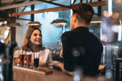 A bartender and a woman engaging in a friendly conversation at a stylish bar, with a flight of craft beers in front of her.