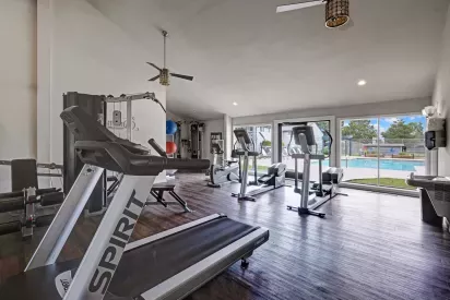 Bright, modern fitness center with treadmills, elliptical machines, weight equipment, and large windows offering a view of the outdoor pool area.