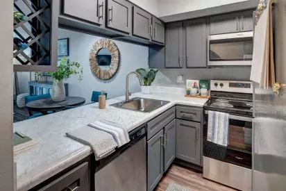 Charming kitchen with modern gray cabinetry, marble-look countertops, and stainless steel appliances. The space includes a sleek undermount sink, electric range, over-the-range microwave, and a pass-through window overlooking the dining area with a round table and contemporary decor.