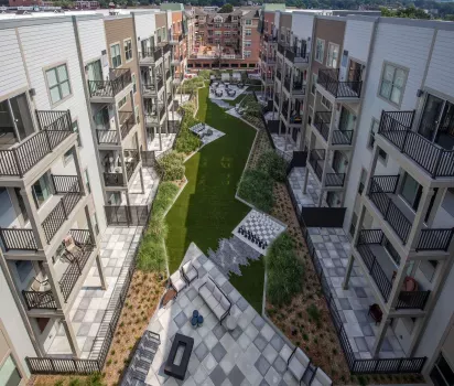 Aerial view of a modern apartment community courtyard featuring a central green space lined with outdoor lounge areas, a life-size chessboard, and firepit seating. The surrounding buildings have private balconies and neutral-toned siding, offering a sleek, contemporary aesthetic. The landscaping includes ornamental grasses and neatly maintained garden beds, creating an inviting atmosphere for residents to relax and socialize.