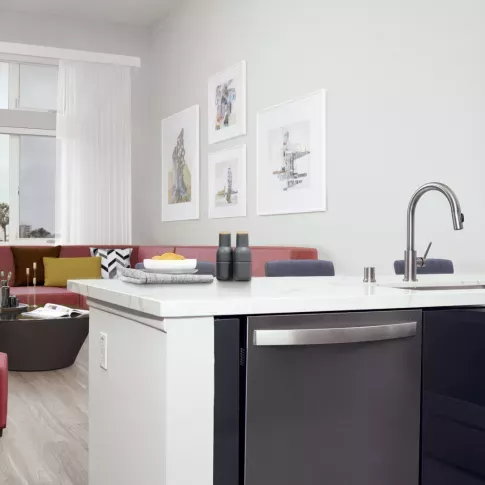 Modern kitchen island with stainless steel appliances and a gooseneck faucet, opening into a stylish living area with a red sectional, geometric ottomans, and framed wall art at The Linden.