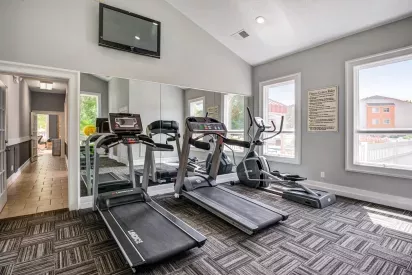 Fully equipped fitness center at The Samuel with treadmills, elliptical machine, wall-mounted TV, and large windows providing natural light