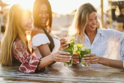 Three young women enjoying refreshing cocktails at an outdoor gathering, toasting with smiles in the warm sunlight.