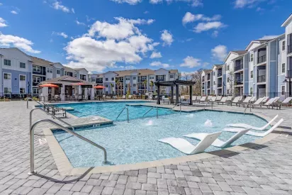 Resort-style swimming pool at Artisan Carolina Forest Apartments in Myrtle Beach, SC, featuring in-water lounge chairs, shaded pergolas, sundeck seating, and modern three-story apartment buildings in the background.