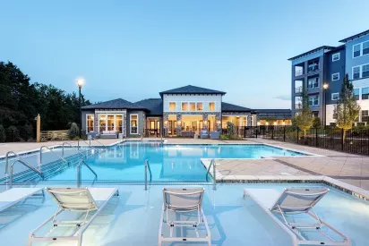 Expansive outdoor swimming pool with built-in lounge chairs on a sun shelf, surrounded by a spacious deck. In the background, a modern clubhouse with large windows glows warmly at dusk, framed by landscaped grounds and contemporary apartment buildings.