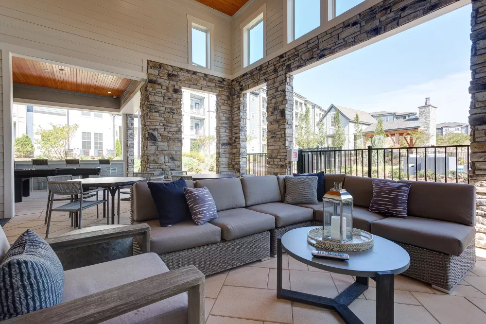Covered outdoor seating area with stone columns, cushioned wicker furniture, accent pillows, and a modern coffee table with lantern décor, overlooking the apartment community.