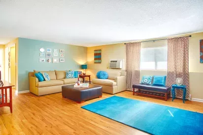 Bright and colorful living room with wood-style flooring, beige sofas, and bold turquoise accents. The space features patterned curtains, a blue accent wall with framed art, a large blue area rug, and decorative pillows. Natural light enters through the window, creating a cheerful and inviting atmosphere.