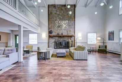 Spacious community lounge with wood-style flooring, high ceilings with exposed beams, and a tall stone fireplace as the focal point. The seating area includes modern sofas, armchairs, and a central ottoman, accented by floor lamps and wall art.