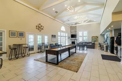 Spacious community clubhouse interior with vaulted ceilings, exposed beams, and large windows letting in natural light. The room features a shuffleboard table on a rug in the center, high-top tables with barstools along one wall, and a lounge area with gray seating, a fireplace, and a wall-mounted TV. The space also includes modern artwork, chandeliers, and a kitchenette with stainless steel appliances.