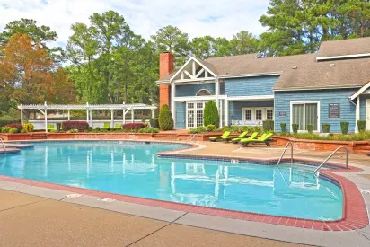 A bright outdoor swimming pool area in front of a blue clubhouse with white trim and a brick chimney. The pool deck features lime green lounge chairs and red brick accents around the water’s edge. Behind the pool, a white pergola with additional seating is surrounded by landscaped greenery and tall trees under a partly cloudy sky.