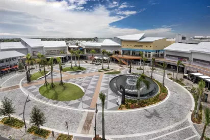 Open-air shopping plaza with palm trees, a large round fountain, and surrounding stores under a bright blue sky.