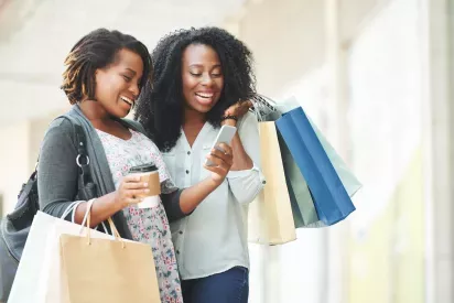 Two smiling women walking outdoors with shopping bags and a coffee cup. One woman is holding a smartphone, and they appear to be laughing and enjoying a conversation. The background is softly blurred, suggesting a bright, pleasant day in a shopping district.