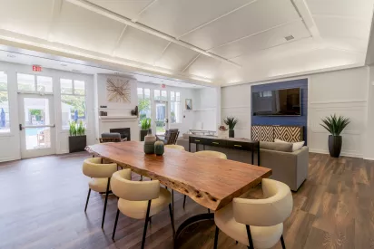 Spacious common room featuring a live-edge wood table, cream chairs, and a sitting area with a TV. A pool is visible through glass doors.